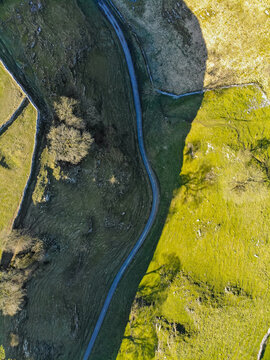 Aerial view of a winding road through green fields and pastures, bordered by dry stone walls and scattered trees under bright sunlight in Buxton, England, United Kingdom.