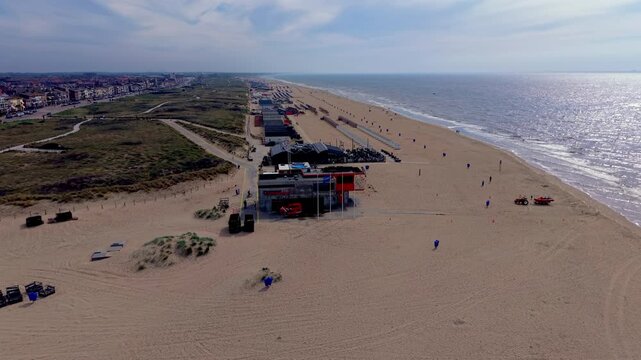 High angle horizontal aerial drone video clip of beach rescue station, restaurant pavilion, cabins, dunes, North Sea waves, and Katwijk aan Zee town skyline, South Holland, Netherlands, West Europe