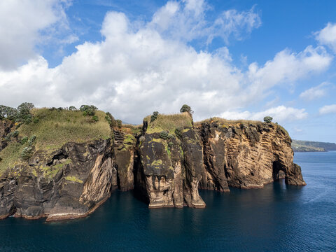 Aerial view of Miradouro do Sertao featuring dramatic volcanic cliffs rising from the deep blue Atlantic Ocean under a bright sky with scattered clouds Capelas, Acores, Portugal.