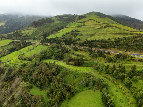 Aerial view of the lush green hills and terraced fields surrounding the Miradouro de Santa Iria with a winding road cutting through the landscape in Porto Formoso, Acores, Portugal.
