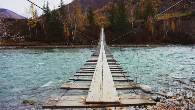 Wooden suspension bridge crosses a turquoise river. Mountains with autumn trees rise in the background. The bridge stretches forward, inviting travelers to cross