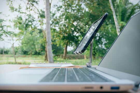 Workspace mockup with laptop, tablet on stand, notebook, and pen on wooden desk with green nature tree background, concept of remote work, work from anywhere, and digital nomad lifestyle.
