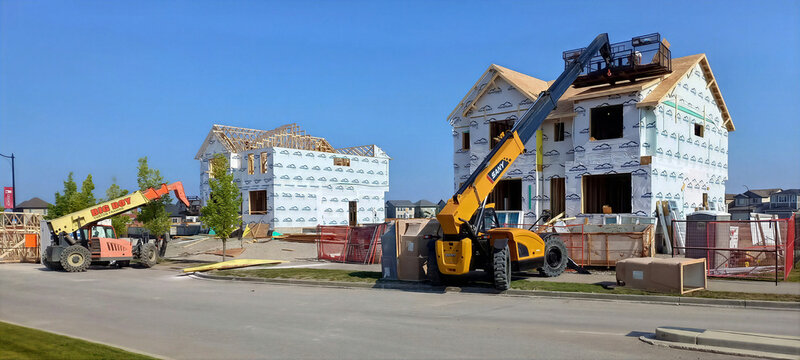 CALGARY, ALBERTA, CANADA - 25 AUGUST, 2023. Two heavy-duty telescopic handlers lift building materials and roofing supplies to the upper floors of unfinished wood-frame houses.