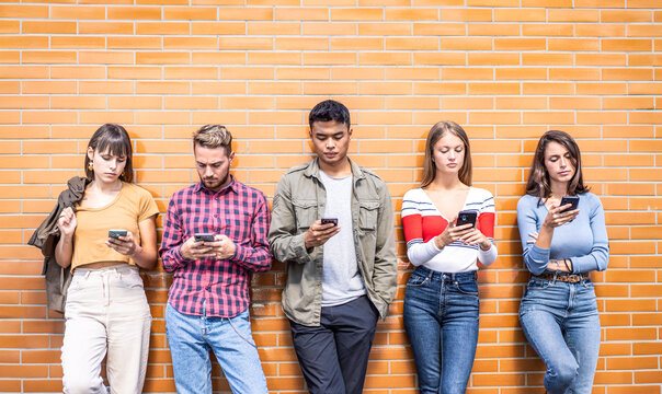 Five gen z friends stand in a row against a brick wall while using smartphones in an urban setting. Front-facing group scene highlights screen addiction, phubbing, and social disconnection.