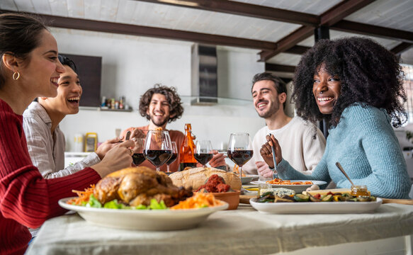 Multicultural young friends laugh together over a homemade dinner with roast chicken and wine at a shared table. Gen z and millennial adults enjoy relaxed conversation and friendship at home.