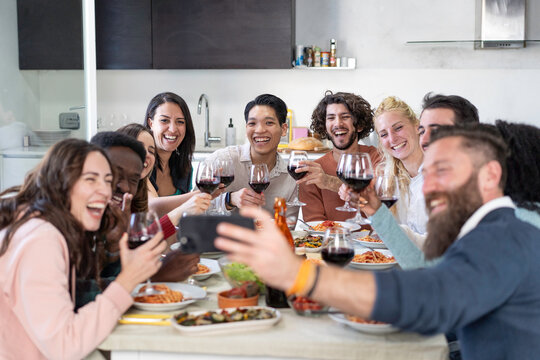 Large diverse group of young friends pose for a smartphone selfie with raised wine glasses during a lively dinner at home. Gen z and millennial adults capture laughter and friendship together.