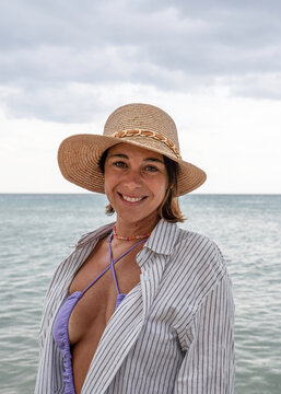 Warm beach smile from a confident gen x woman in a straw hat by the sea. Vertical lifestyle portrait with direct eye contact and copy space, ideal for wellness, travel and book cover use.