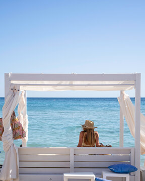 Framed by a white beach cabana, a millennial woman sits facing the turquoise sea on a sunny summer vacation. Vertical resort scene with blue sky and copy space for travel and book cover use.