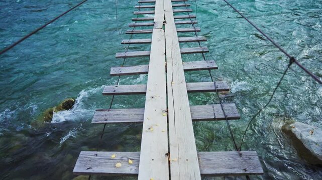 Rusty wooden planks form a narrow suspension bridge over rushing turquoise water. Dark cables stretch taut, holding the structure above churning currents. Stones peek from clear, cold depths below