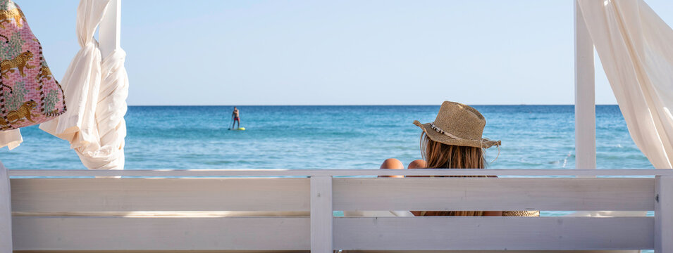 Panoramic resort banner showing a woman relaxing under a white cabana facing the turquoise sea. Wide beach header with copy space, ideal for travel, hotel and hospitality marketing.
