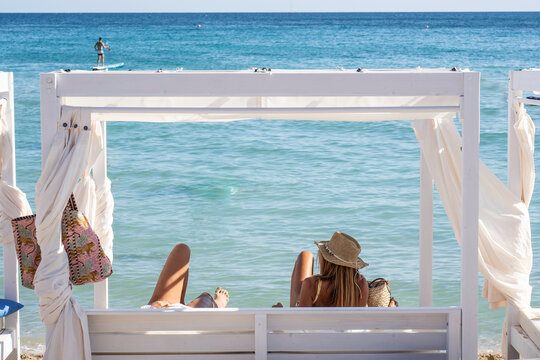 Two women unwind beneath a white seaside cabana during a sunny beach vacation. One faces the turquoise sea while her companion reclines beside her. Shared leisure and summer escape concept.