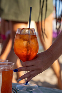 A chilled orange spritz cocktail with citrus and ice is held at a lively summer beach bar. Close-up vertical lifestyle scene with condensation.