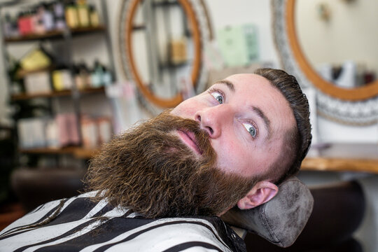 Bearded millennial man reclines in a barber chair while showing his long groomed beard in a modern salon. Close-up portrait conveys masculine style, relaxation, and playful confidence.