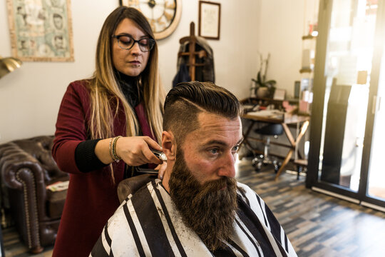 Female millennial barber uses electric clippers to refine a bearded client's fade and sideburns in a modern salon. Authentic grooming scene highlights precision, style, and professional care.