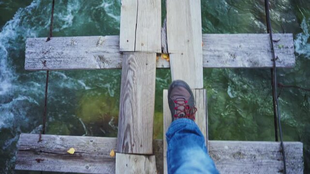Hiker steps carefully across a rustic wooden bridge over rushing green water. The camera focuses on one foot, highlighting the tension of crossing. Water splashes beneath