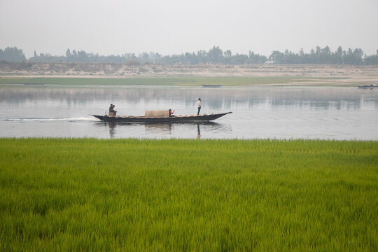 Bogura, Rajshahi Division, Bangladesh - 28 February 2022: View of a wooden boat carrying people across a calm river with lush green fields in the foreground under a hazy sky.