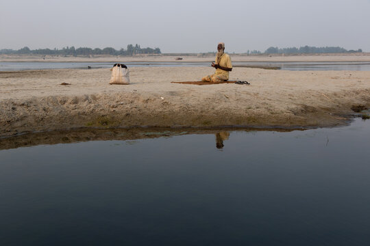 Bogura, Rajshahi Division, Bangladesh - 28 February 2022: View of an elderly man sitting on a prayer mat on the sandy bank of the Paryer dry riverbed.