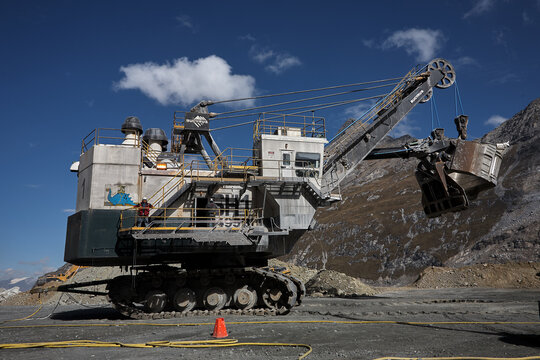 An electric rope shovel stands ready at an open-pit copper site, its massive dipper arm raised against a dramatic backdrop of terraced rock benches carved deep into the earth