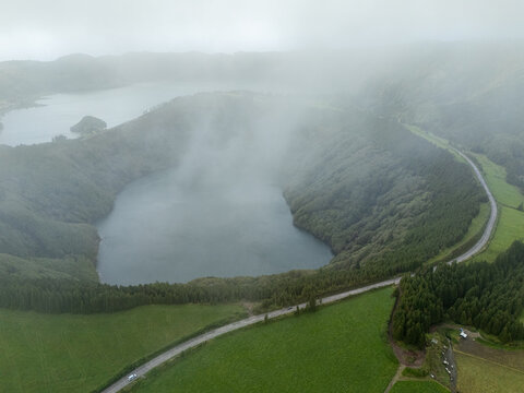 Aerial view of a volcanic crater lake surrounded by lush green hills, dense forests, and a winding road partially obscured by thick mist, Azores, Portugal.