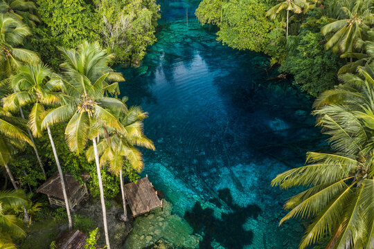 Aerial view of Danau Paisu Pok Luk Panenteng, a crystal clear blue lake surrounded by lush palm trees and wooden structures in Bulagi Utara, Central Sulawesi, Indonesia.