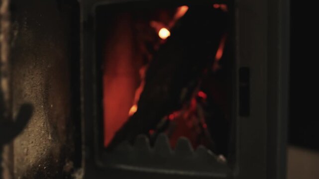 First-person view of a person opening a metal wood-burning stove with glowing firewood inside.