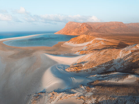 Aerial view of Detwah Lagoon Ramsar Site with white sand dunes, turquoise waters, and rocky mountains under a clear sky in Socotra, Yemen.