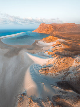 Aerial view of Detwah Lagoon Ramsar Site featuring white sand dunes, turquoise waters, and rugged mountains under soft golden light in Socotra, Yemen.