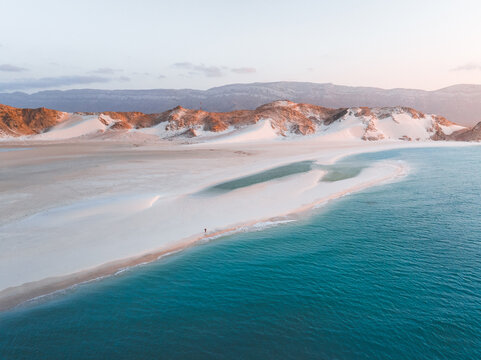 Aerial view of Detwah Lagoon Ramsar Site with white sand dunes and turquoise water against a backdrop of mountains in Socotra, Yemen.