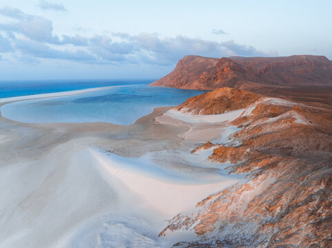 Aerial view of Detwah Lagoon Ramsar Site with white sand dunes, turquoise waters, and rocky cliffs under golden hour light in Socotra, Yemen.