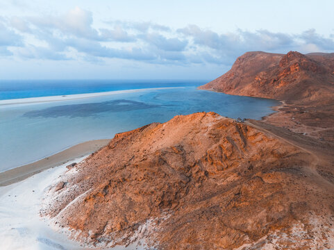 Aerial view of Detwah Lagoon Ramsar Site with its white sandbar, turquoise waters, and rocky hills under a soft sky in Socotra, Yemen.
