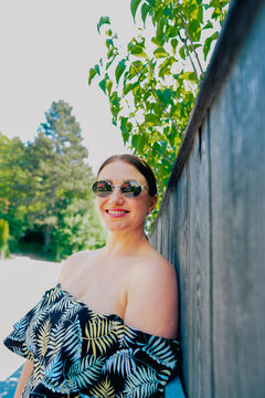 Woman with brown hair wearing sunglasses and a tropical-patterned off-shoulder top leans against a wooden fence with greenery in the background on a sunny day