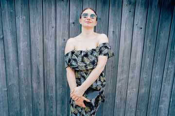 Fototapeta premium Female model wearing off-shoulder tropical print dress poses confidently against a wooden wall, showcasing summer fashion with sunglasses and a clutch bag