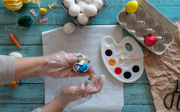 Creative Easter craft scene showing hands painting colorful eggs with brushes and a watercolor palette surrounded by additional eggs and craft supplies on a rustic tabletop.