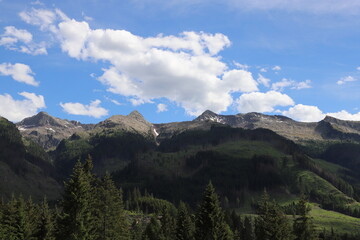 Obraz premium Panoramic view of forested valleys and distant mountain ranges beneath a sky filled with dramatic cumulus clouds in the Italian Alps during summer.