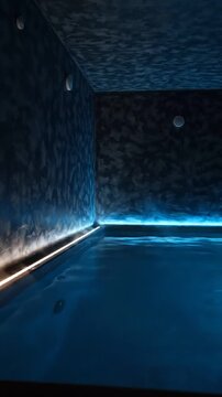 Indoor swimming pool with illuminated edges transitioning from warm to cool lighting, showcasing reflections on water surface in a modern, minimalist setting