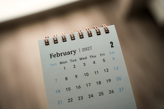 Small spiral desk calendar showing February 2027 dates on a wooden table with dramatic side lighting