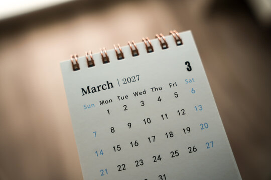 Small spiral desk calendar showing March 2027 dates on a wooden table with dramatic side lighting