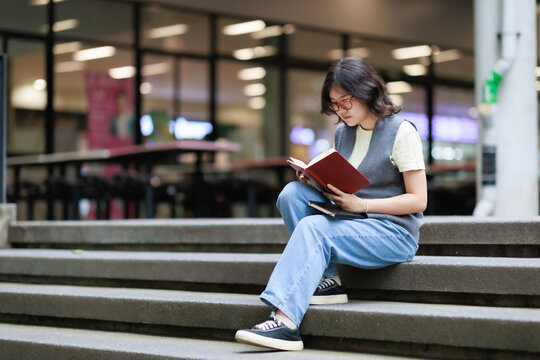 Young female student with short dark hair and glasses sitting on campus stairs reading a book, focusing on education and academic life outdoors