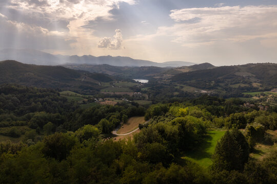 Aerial view of the rolling hills and lush forests surrounding the village of Smerillo, with sunbeams breaking through clouds over the distant mountains in Smerillo, Marche, Italy.
