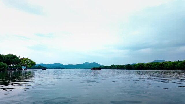 A tranquil lake scene with gentle ripples, flanked by dense green trees and distant blue mountains under an overcast sky, with a small boat on the water.
