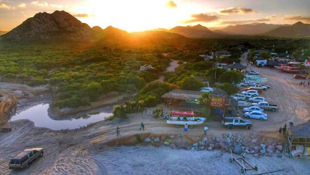 Aerial view of Parque Nacional Cabo Pulmo at sunset with desert mountains, coastal vegetation, parked vehicles, and small boats along the shore in Cabo Pulmo, Baja California Sur, Mexico.