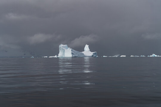 Aerial view of a large white iceberg floating in the dark waters of the Southern Ocean under a dramatic overcast sky in Antarctica.