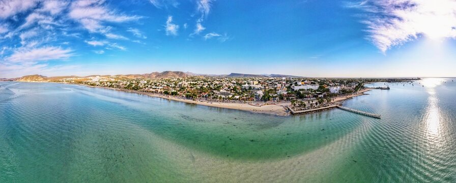 Aerial view of the coastal city with its turquoise waters, sandy beach, palm-lined promenade, and urban buildings under a clear blue sky in La Paz, Baja California Sur, Mexico.