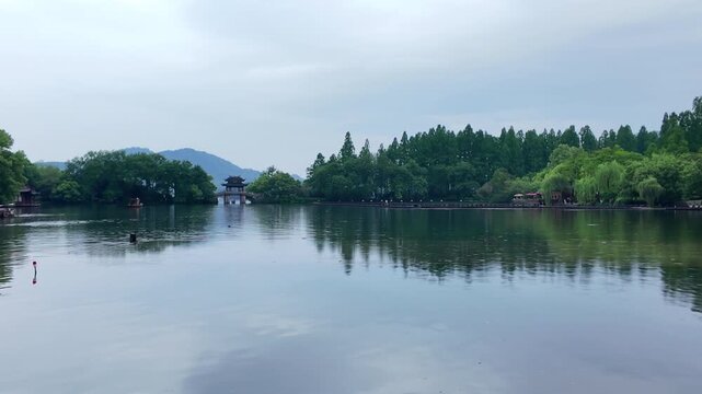 A serene lake landscape with lush trees, distant mountains, and a traditional pavilion reflected in calm water under an overcast sky.