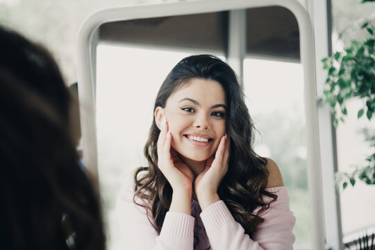 Young woman smiling at mirror while touching her face, confident self care and natural beauty portrait in bright indoor setting