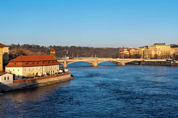 Naklejka premium 10th January, 2024, Czech Republic. A wide river flows under a stone bridge with buildings on the far bank and a red-roofed building on the near bank.