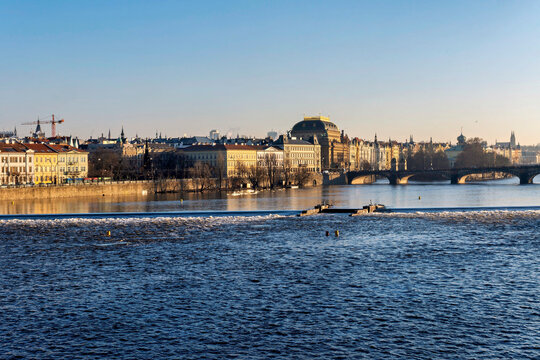 10th January, 2024, Czech Republic. A wide shot of the Vltava River in Prague, Czech Republic, with the National Theatre and other historic buildings lining the riverbank under a clear blue sky.