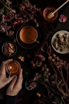 Woman hands holding bowl with honey beside cup of herbal tea and dried flowers on dark velvet fabric.