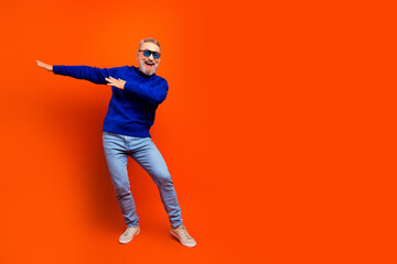 Happy senior man dancing and posing in a blue sweater and sunglasses against a vibrant orange...