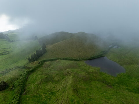 Aerial view of Lagoa Pau Pique surrounded by lush green rolling hills and volcanic landscape under a misty sky in Capelas, Azores, Portugal.
